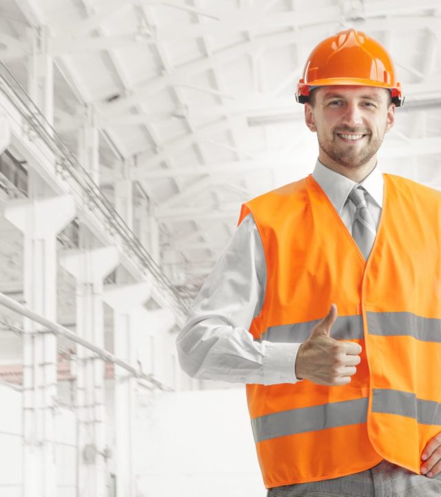 The builder in orange helmet against industrial background The builder in a construction vest and orange helmet smiling with sign ok against industrial background. Safety specialist, engineer, industry, architecture, manager, occupation, businessman, job concept
